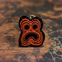 Front of the pin, which is a stone tiki face rendered in black metal and red enamel.